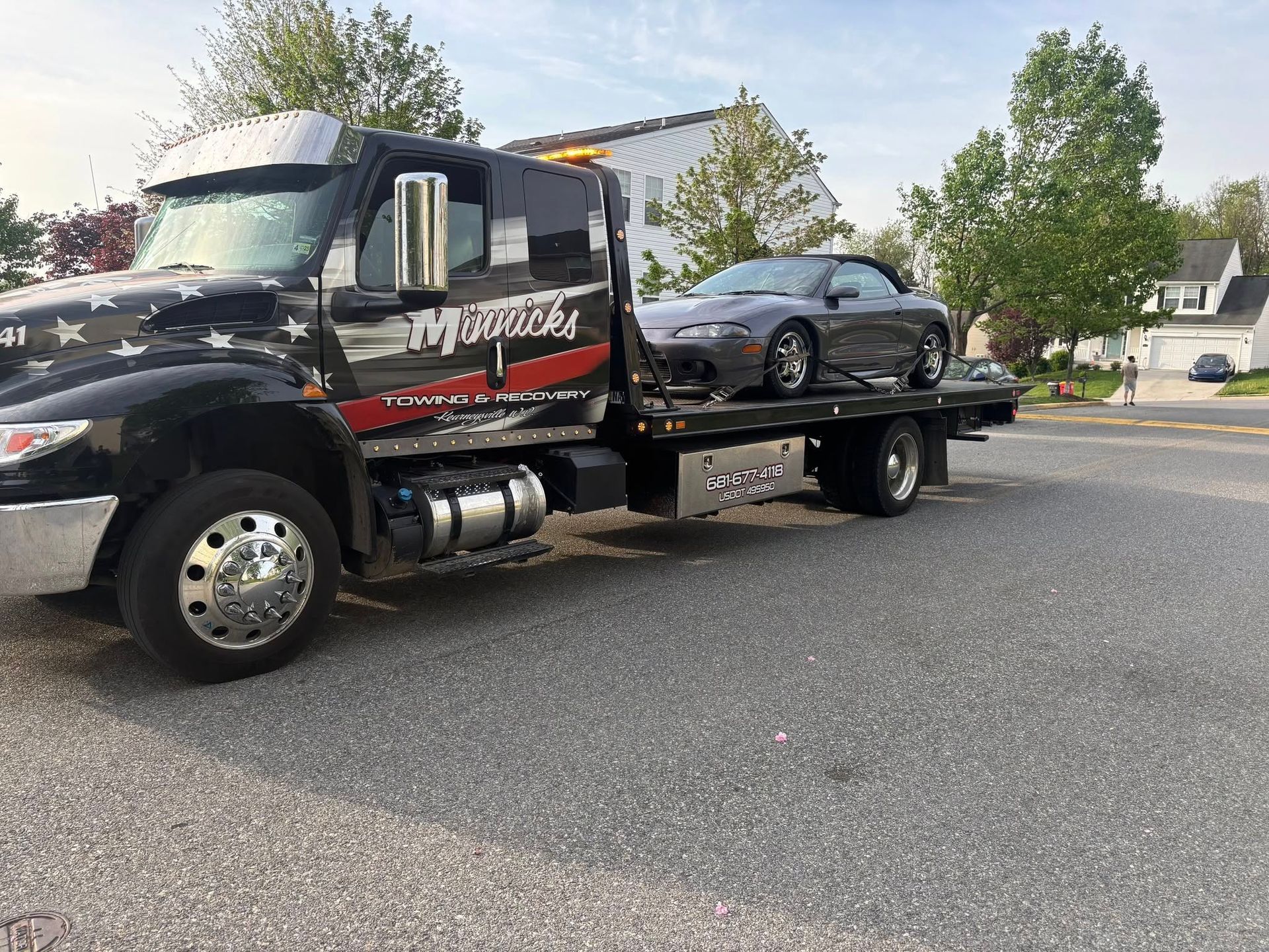 Tow truck with a gray convertible car loaded on its flatbed, parked on a street in front of houses.