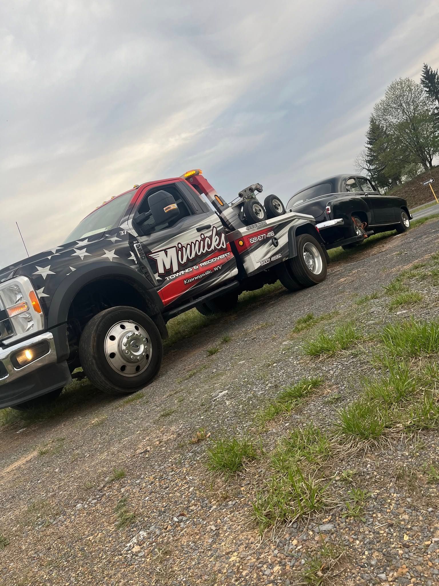 Tow truck with patriotic design towing a classic black car on a gravel road under a cloudy sky.