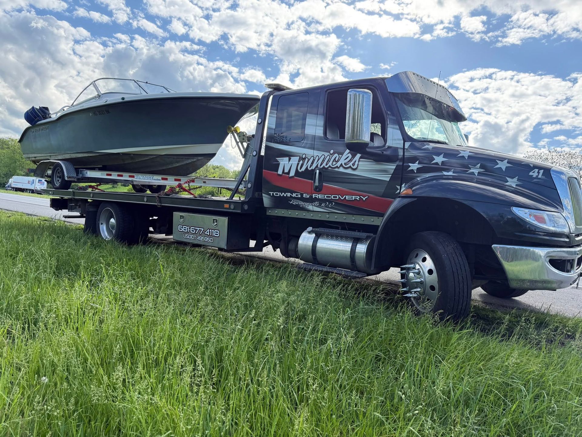 Tow truck with a boat on a trailer, parked on the side of a grassy road, under a cloudy sky.