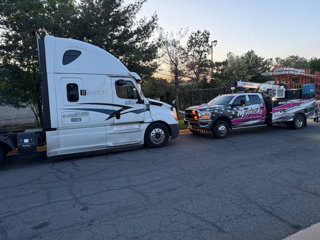 White semi-truck and a pickup truck with a crane are parked side by side on a paved area, trees in the background.