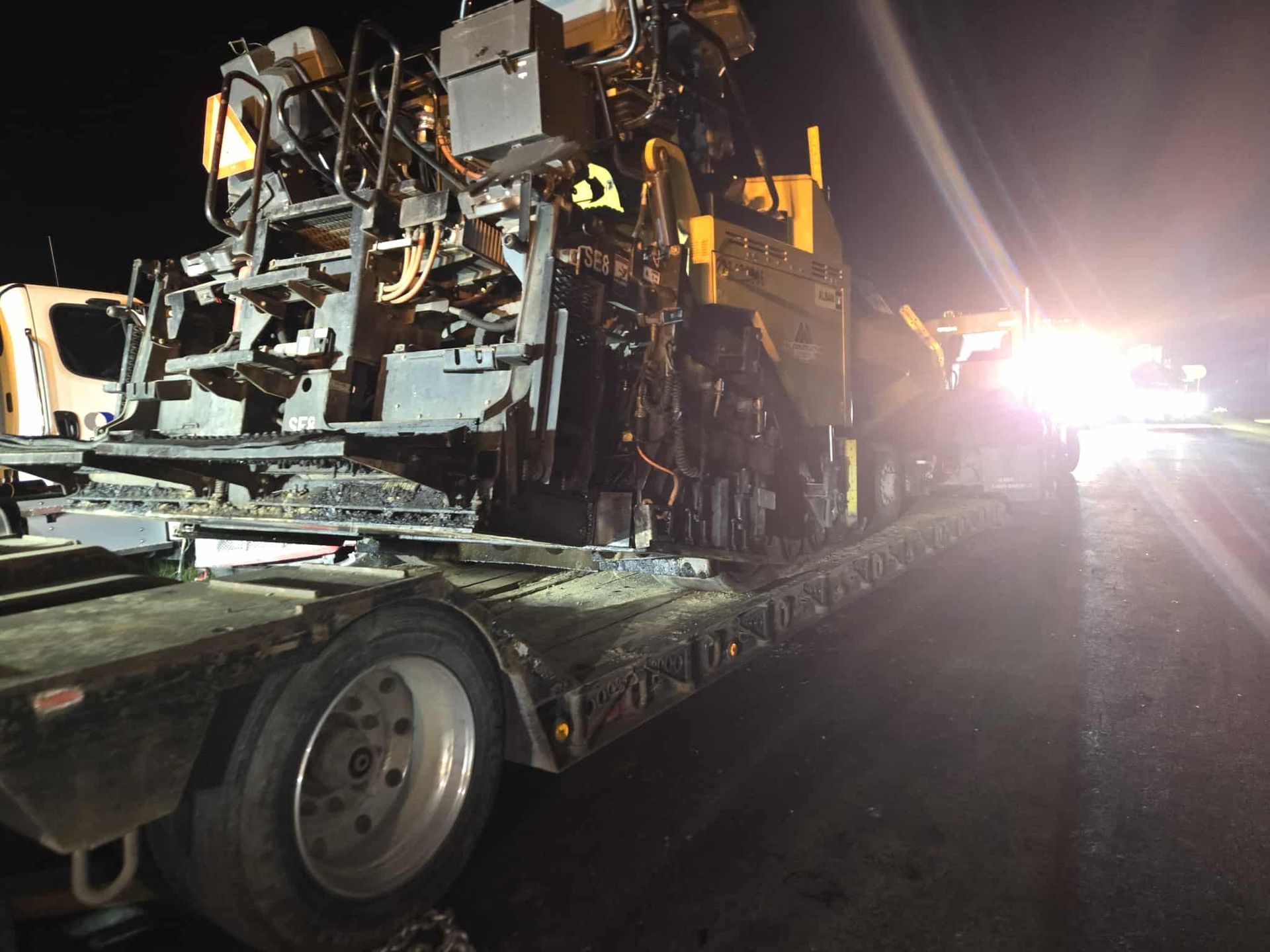 Asphalt paving machine on a flatbed trailer at night, headlights illuminating the scene.
