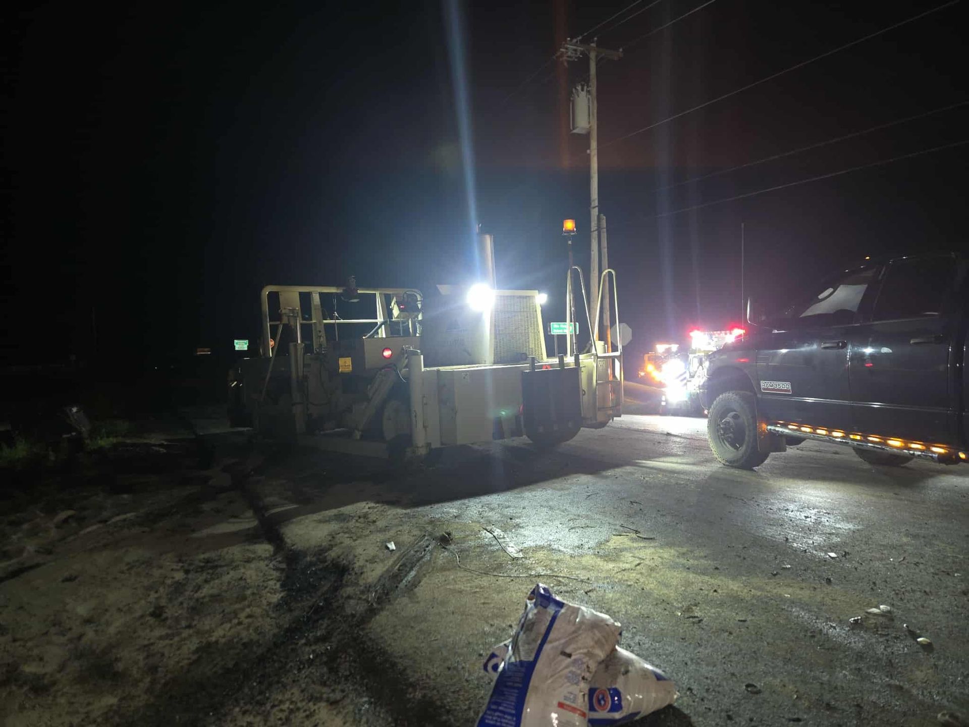 A large construction machine and a dark pickup truck are lit up at night. Debris is on the ground.