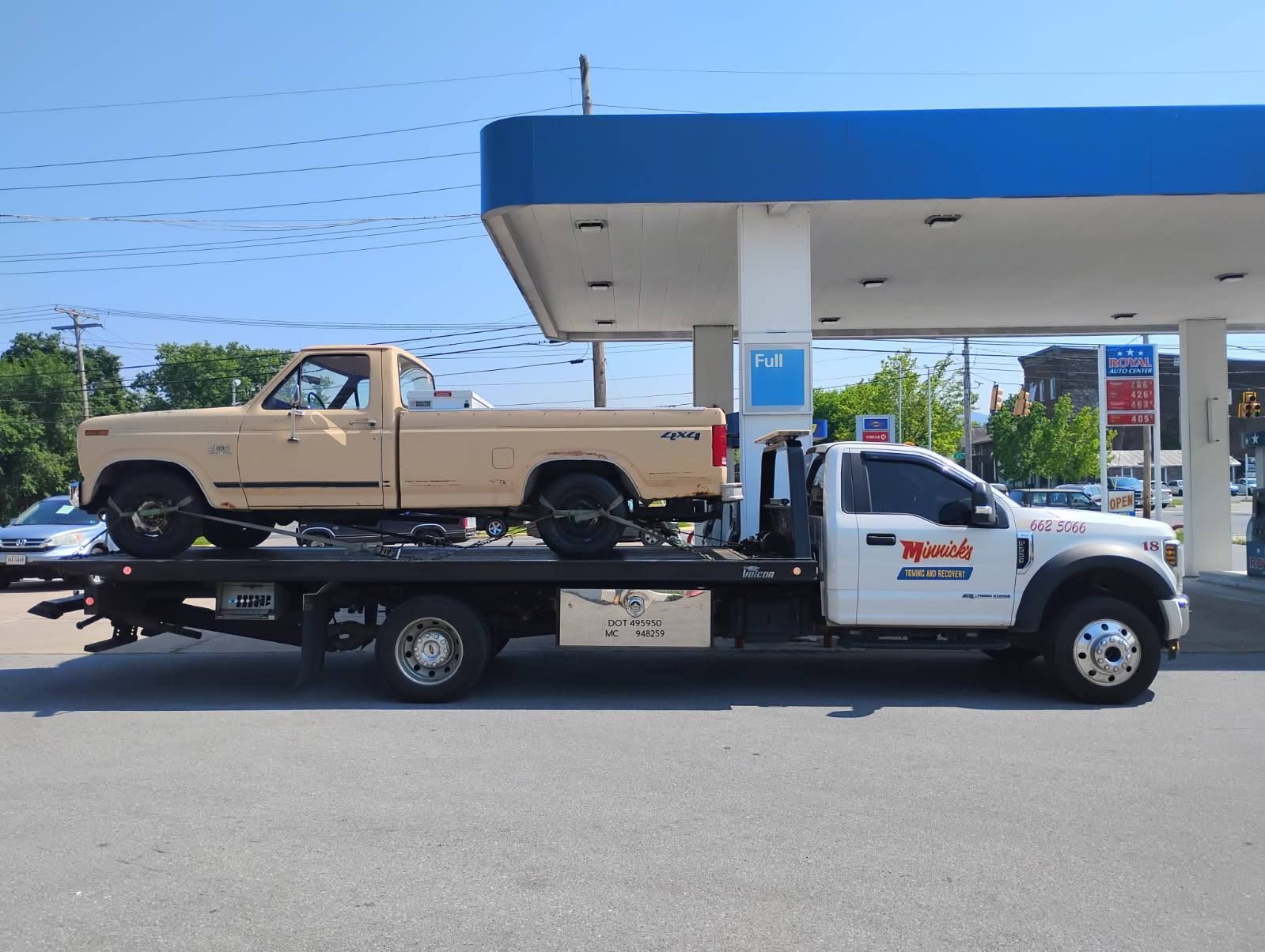 A beige pickup truck being towed by a white tow truck at a gas station under a blue canopy.