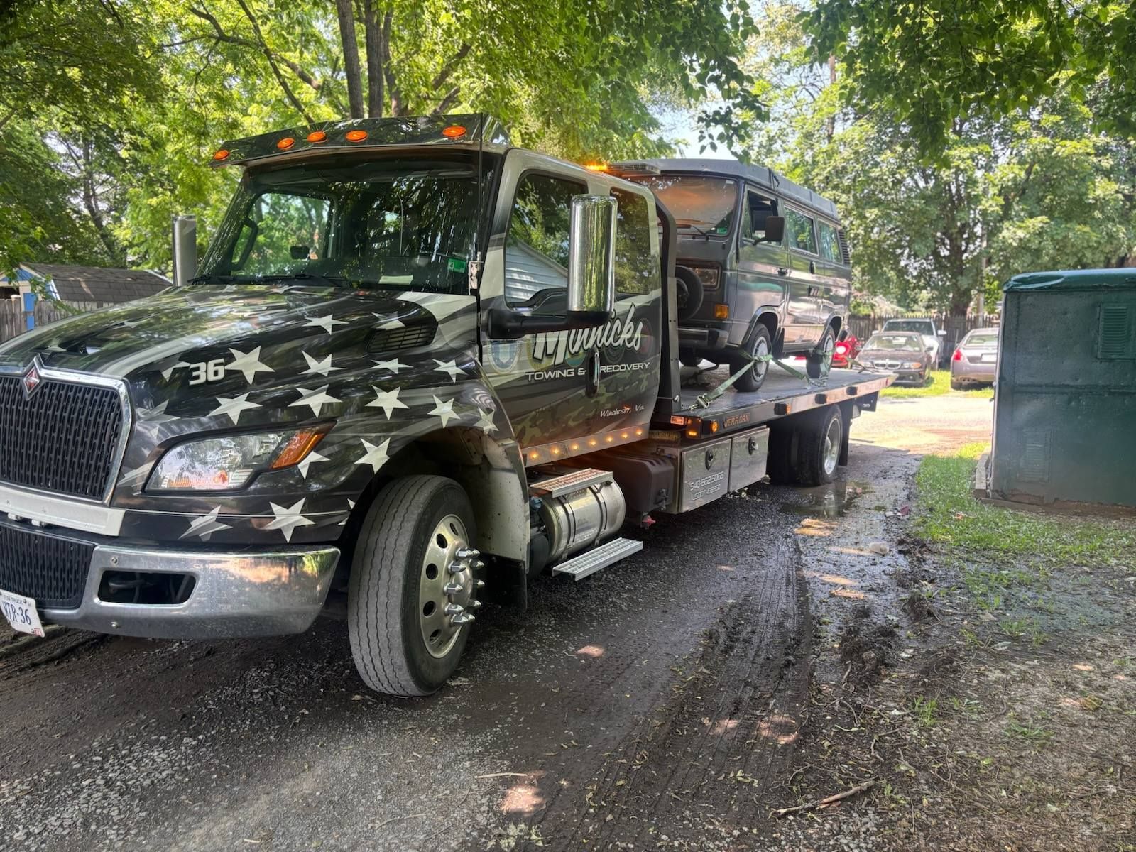 Tow truck with a van loaded on the flatbed, parked on a muddy driveway, next to a green dumpster.