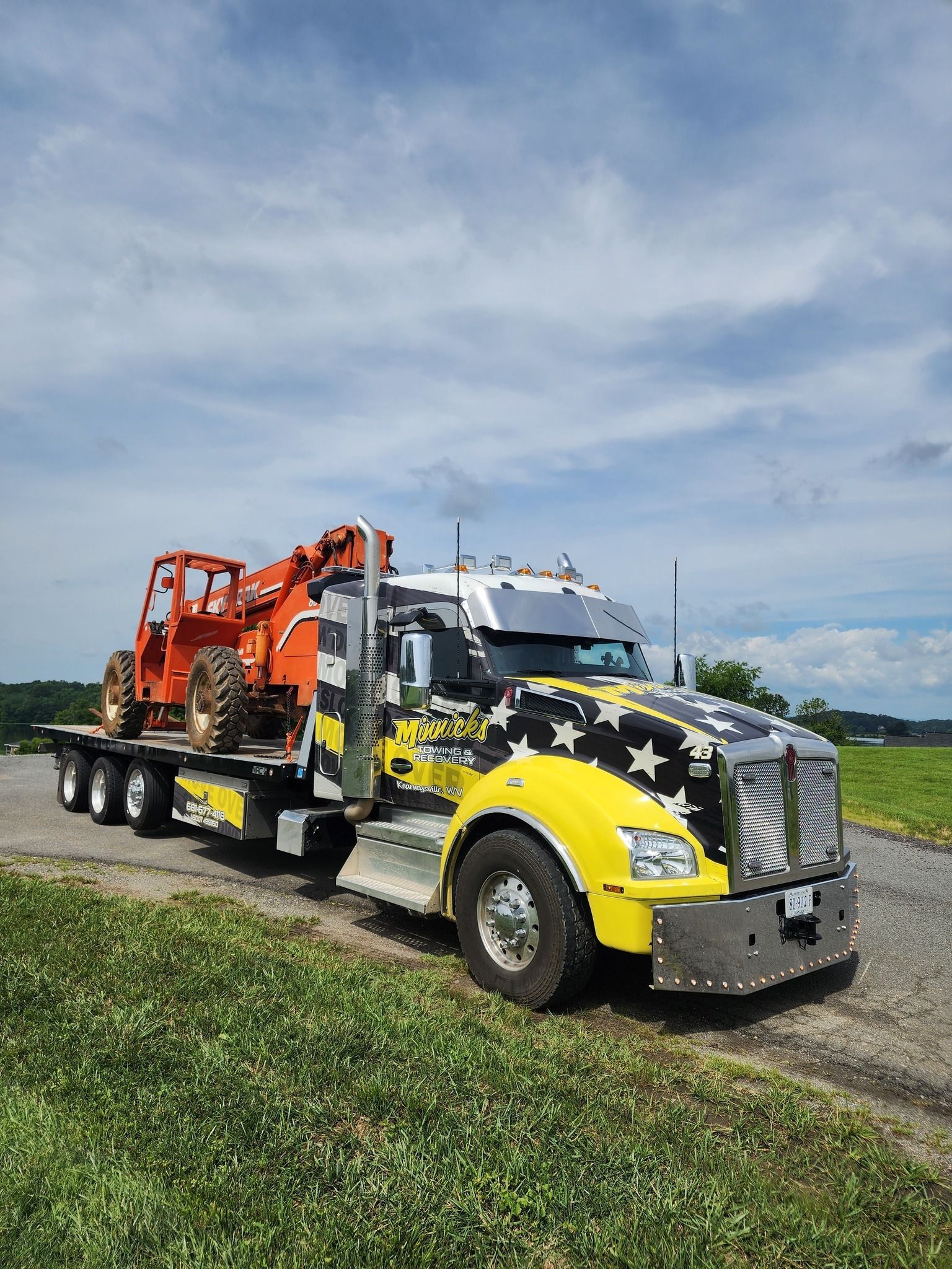 Truck hauling an orange construction vehicle on a flatbed trailer, yellow and star-patterned cab, parked on grass.