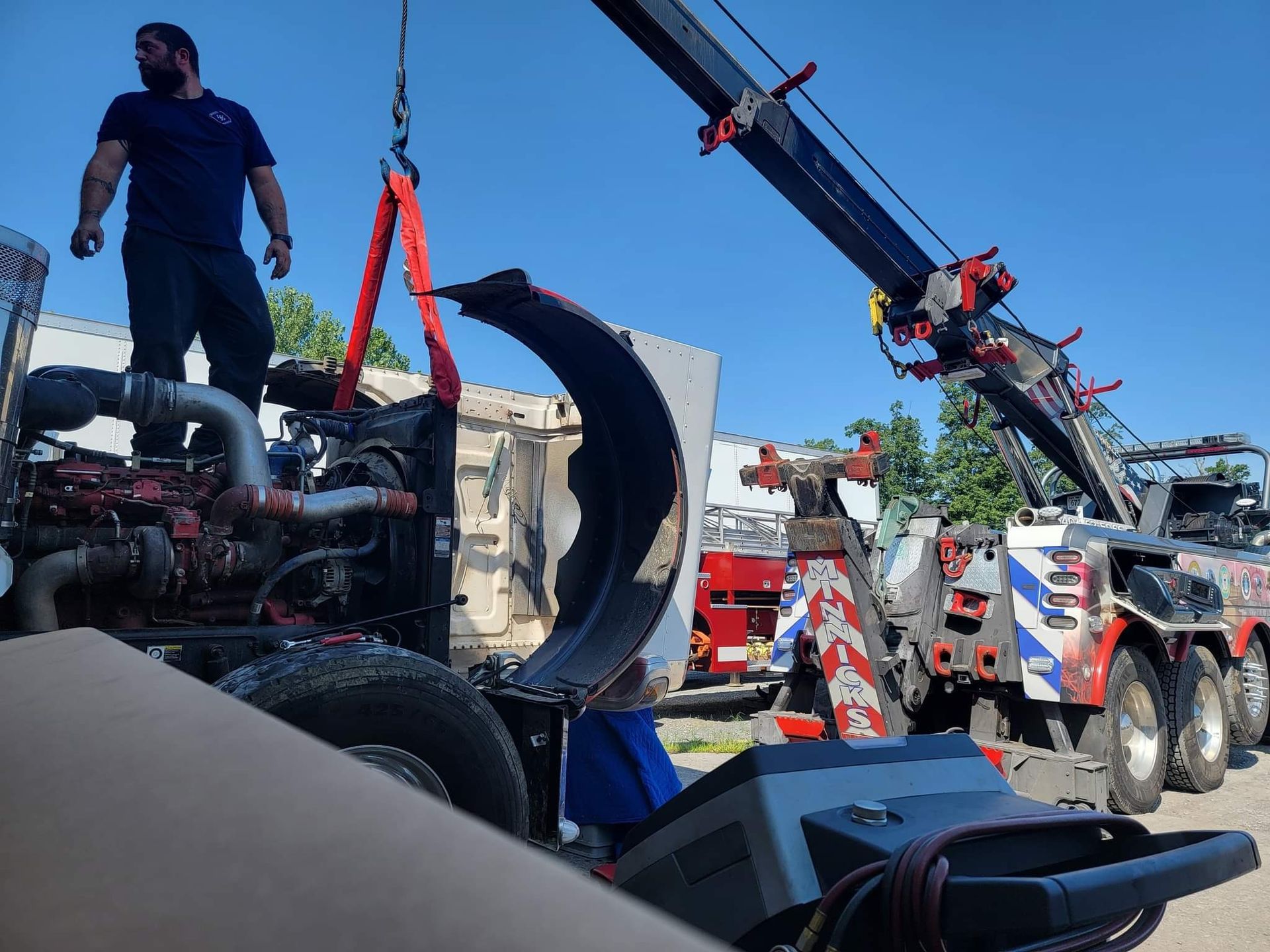 A tow truck lifts a large truck part, another man stands on the truck under blue sky.