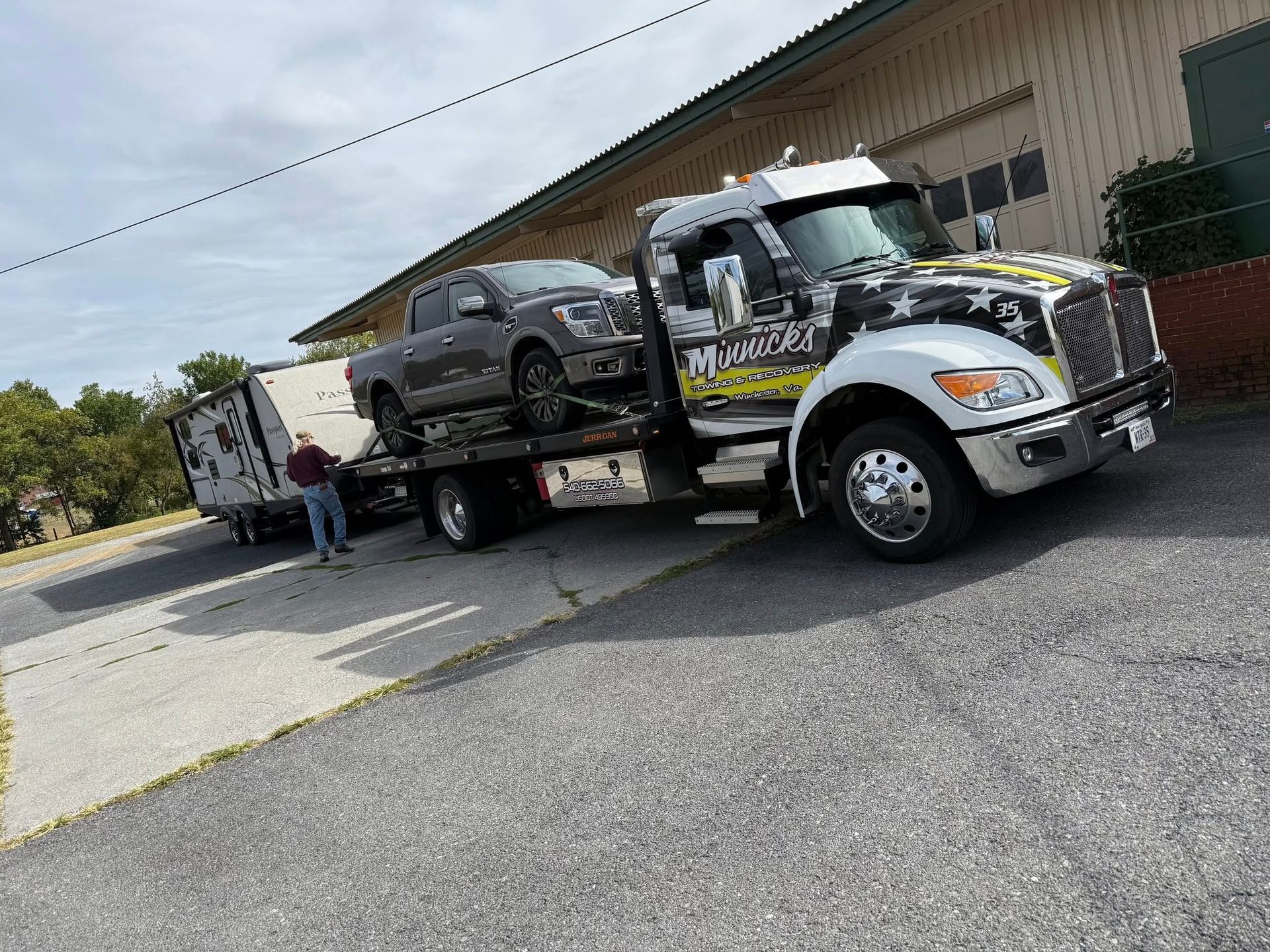 Tow truck hauling a gray pickup truck and trailer on a sloped driveway next to a building.