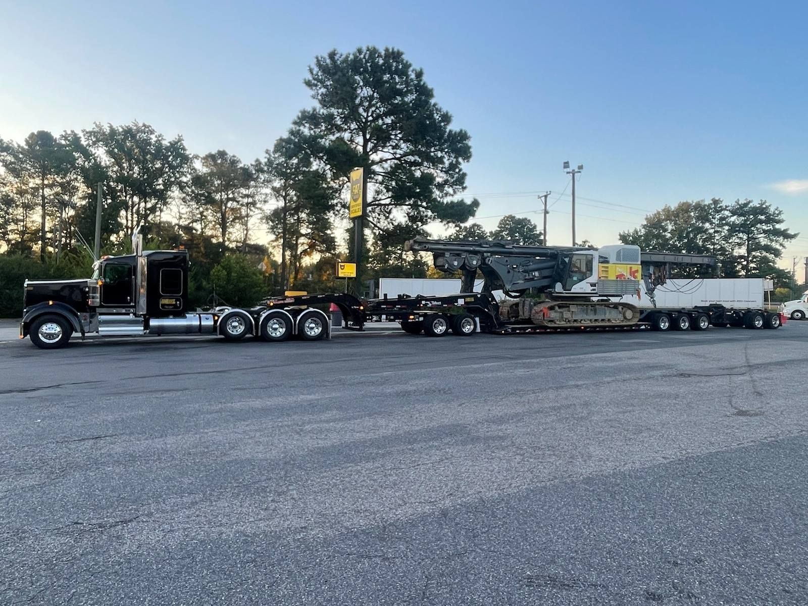 Black semi-truck hauling large construction equipment on a flatbed trailer in a parking lot.