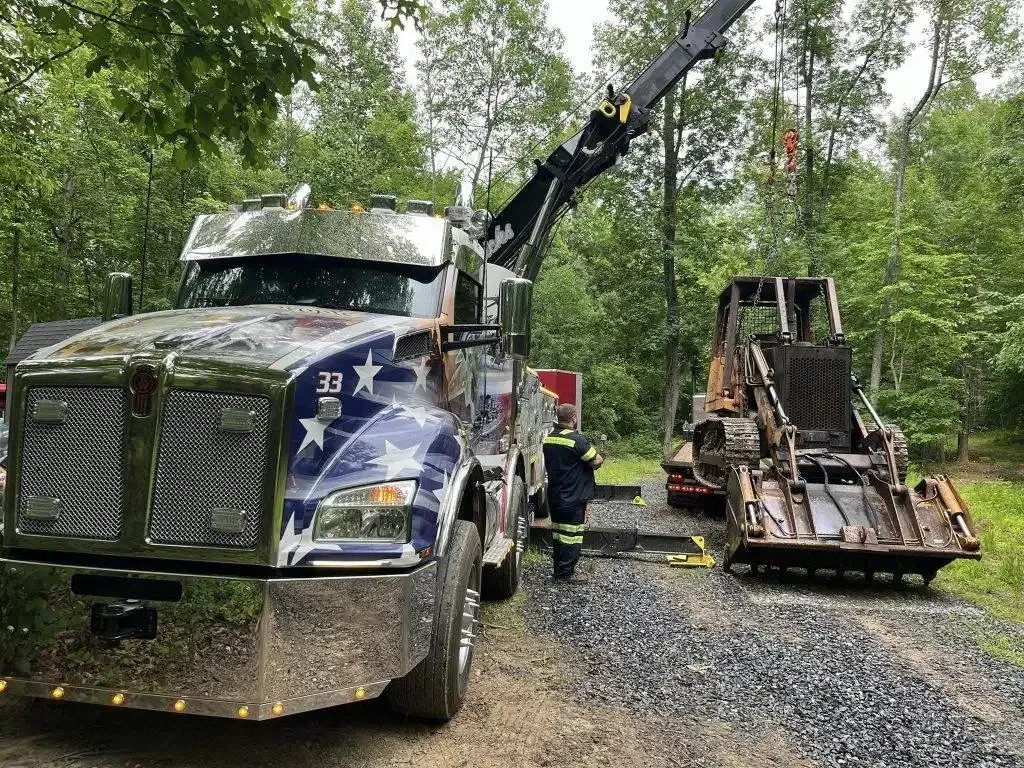 Tow truck lifting a damaged bulldozer in a wooded area. A firefighter stands by.