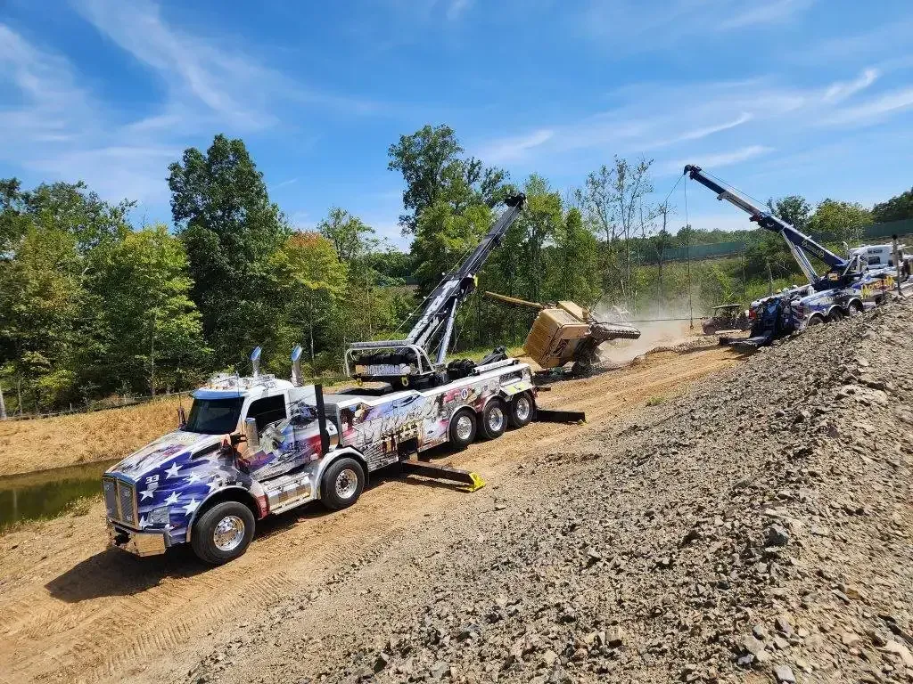 Two tow trucks removing an overturned vehicle from a ditch on a dirt road.
