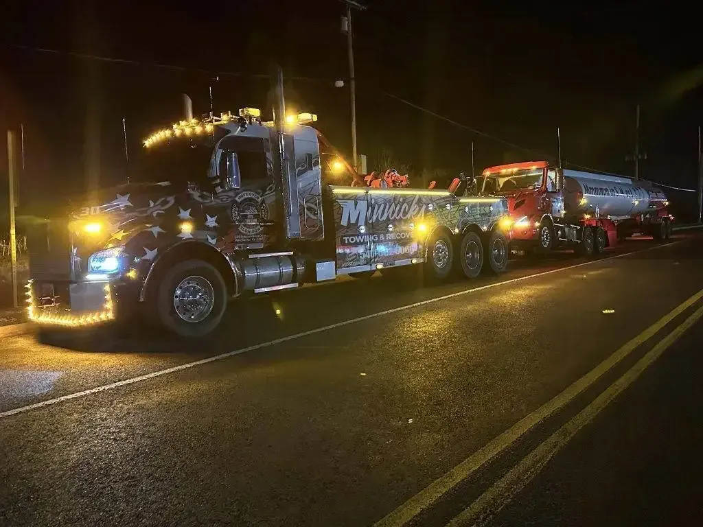 A decorated tow truck with a smaller truck and tanker, illuminated on a dark road.