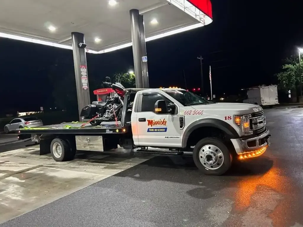 Tow truck with a motorcycle on its bed at a gas station at night. Truck is white with orange lights.