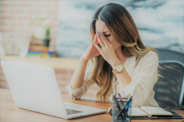 A woman is sitting at a desk with a laptop and covering her face with her hands.