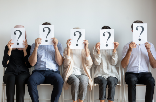 A group of people are sitting in chairs holding signs with question marks on them in front of their faces.