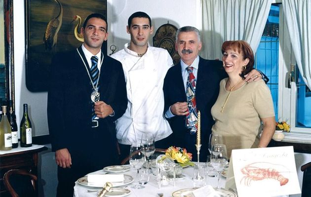 Quattro persone sedute al tavolo di un ristorante: due uomini in giacca e cravatta, uno chef e una donna sorridente.