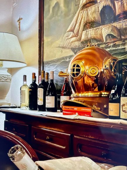 Bottiglie di vino su una credenza in legno, con un casco da sub, sullo sfondo un dipinto raffigurante una nave.