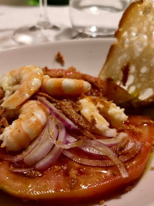 Gamberi, cipolla rossa e pane tostato su fette di pomodoro, in primo piano su un piatto bianco.