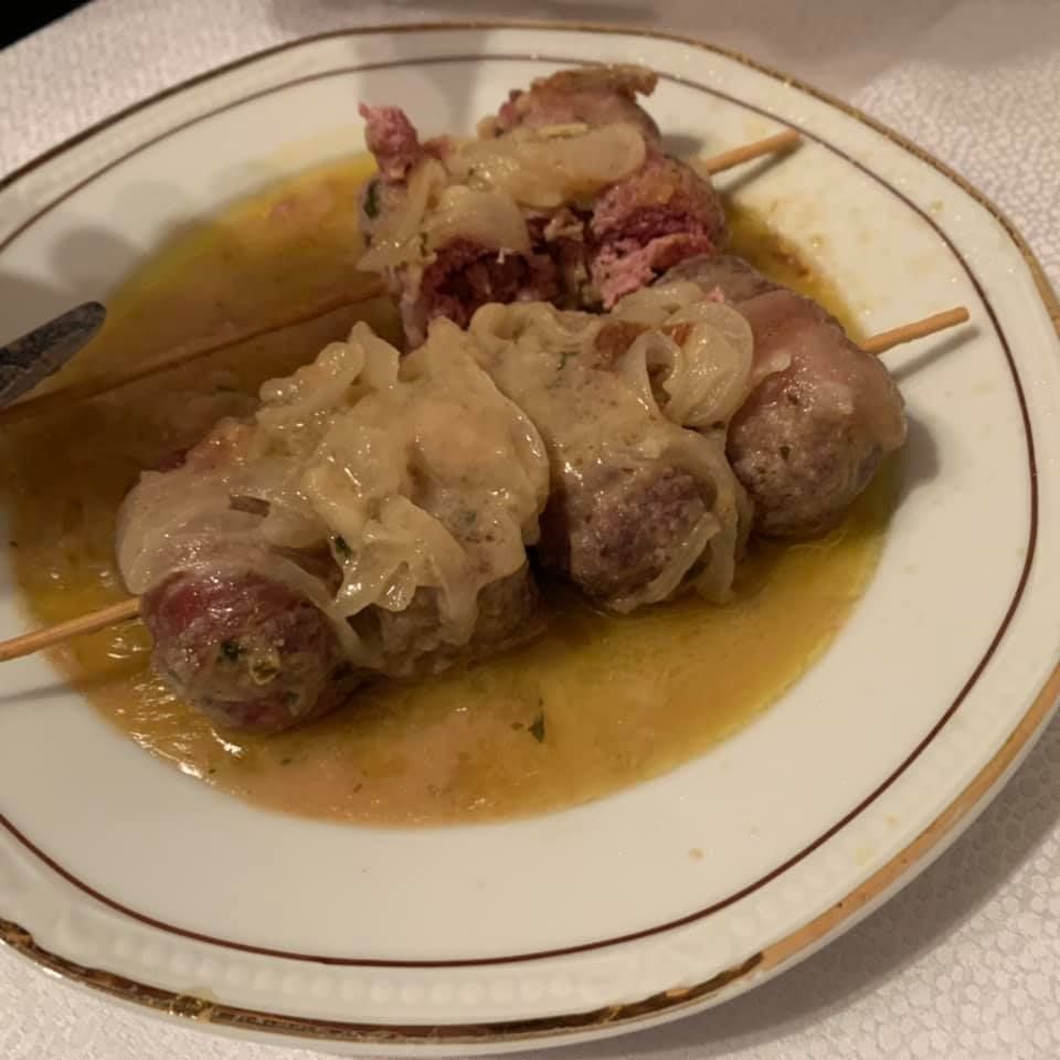 Polpette infilzate con cipolle in salsa su un piatto bianco con bordo dorato.