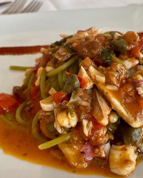 Pasta verde condita con sugo di pomodoro ai frutti di mare, capperi e un filo di salsa rossa su un piatto bianco.