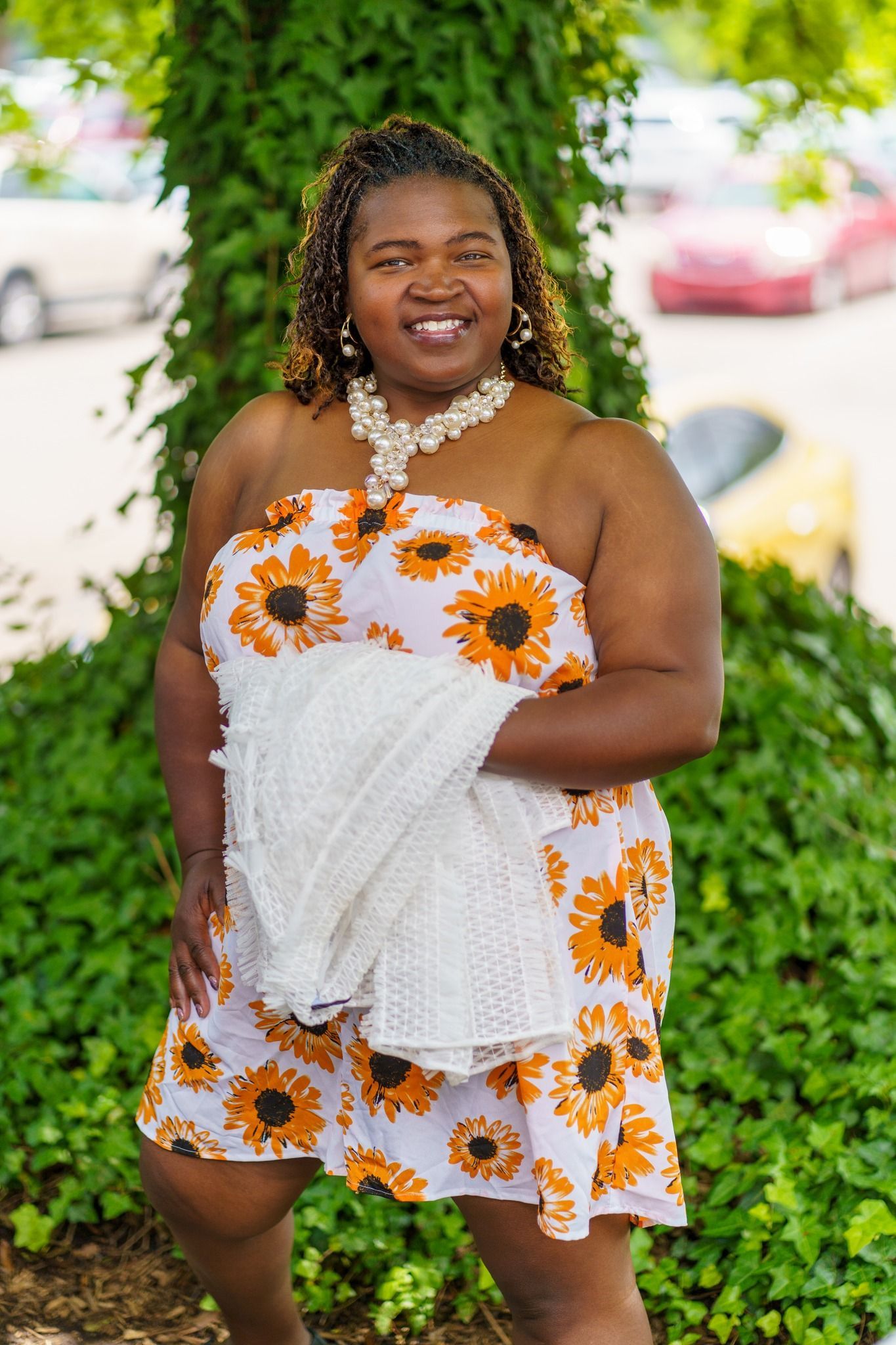 A woman in a sunflower dress is standing in front of a tree holding a white shirt.