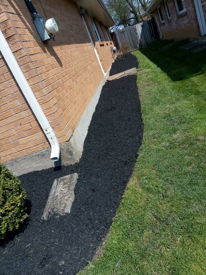 A freshly mulched garden bed borders the brick exterior wall of a house, next to a green lawn.