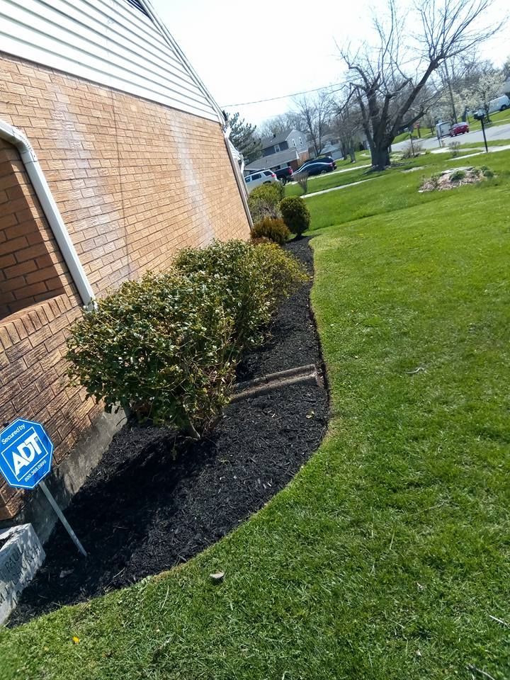 A brick house corner with a curved garden bed containing shrubs and black mulch next to a green lawn.