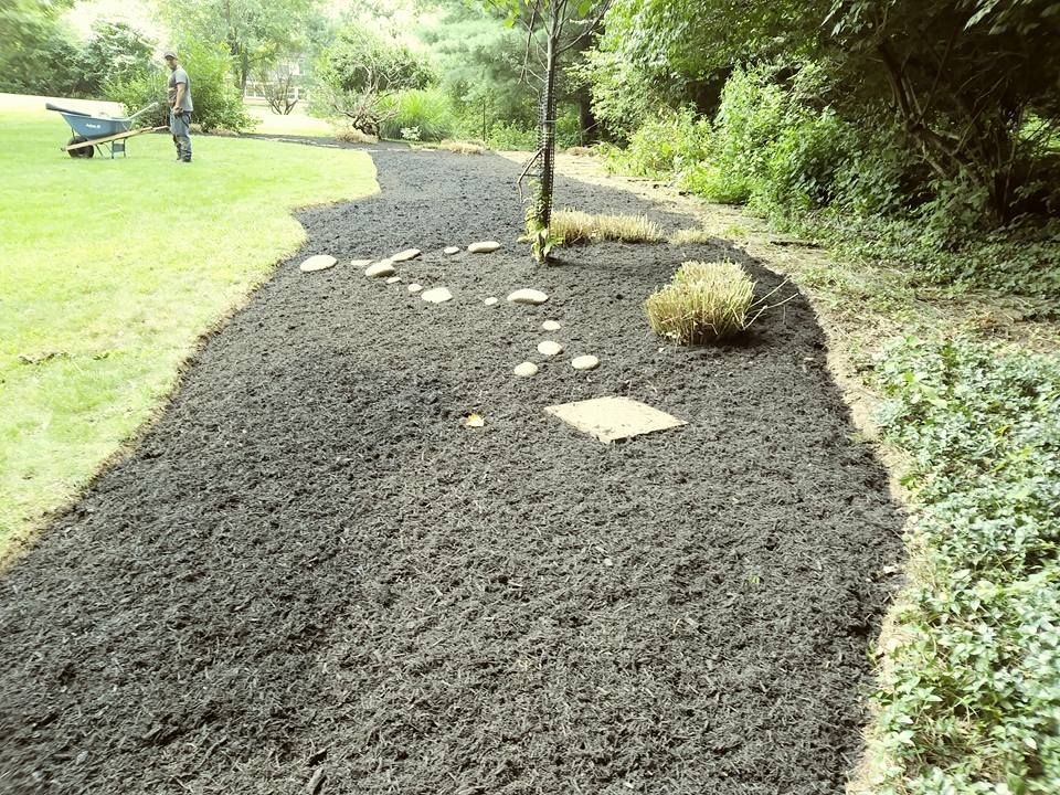 A person pushes a wheelbarrow along a freshly mulched garden bed with stone pavers in a grassy outdoor setting.