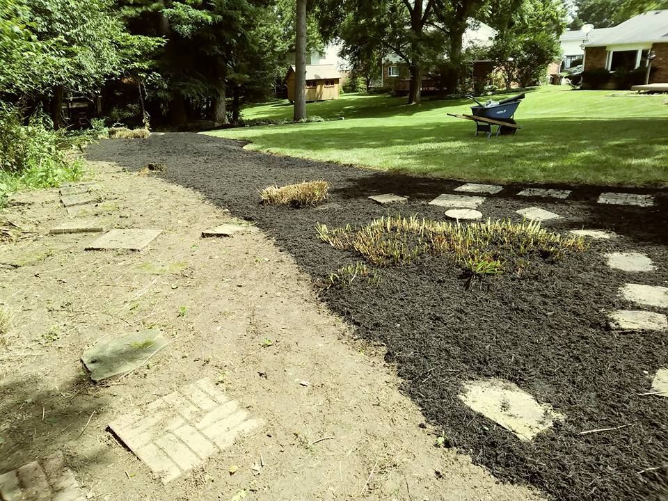 A backyard project showing a transition from bare dirt to a new mulch garden bed with stone pavers laid out in a path.