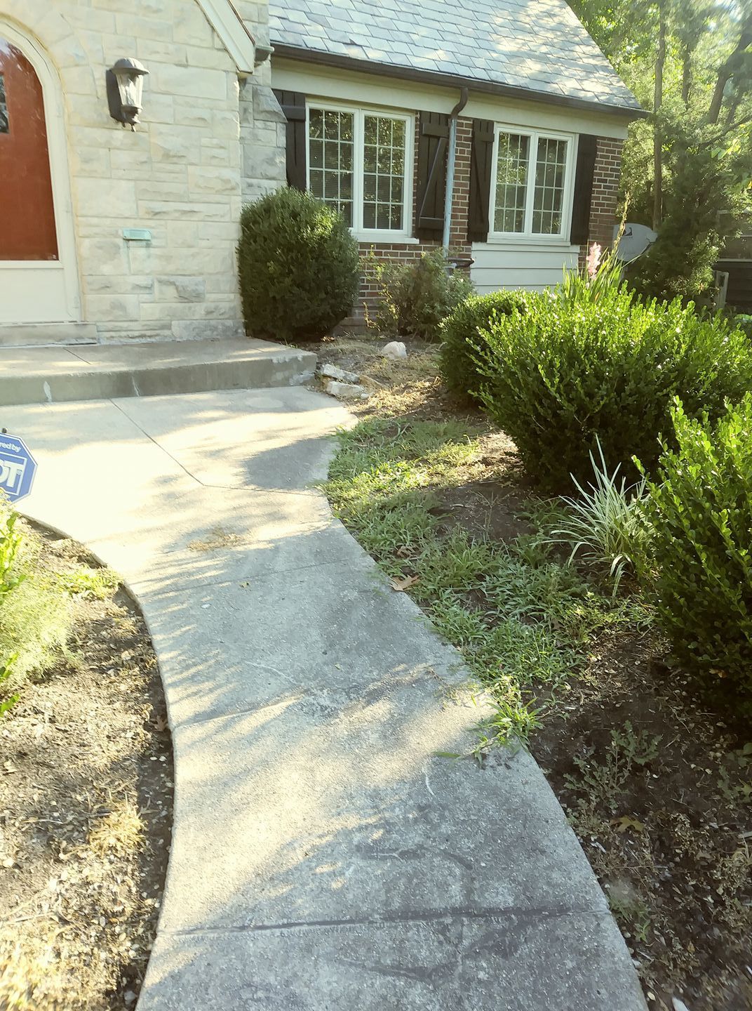 A concrete walkway leads toward the entrance of a stone house with dark shutters and surrounding green landscaping.