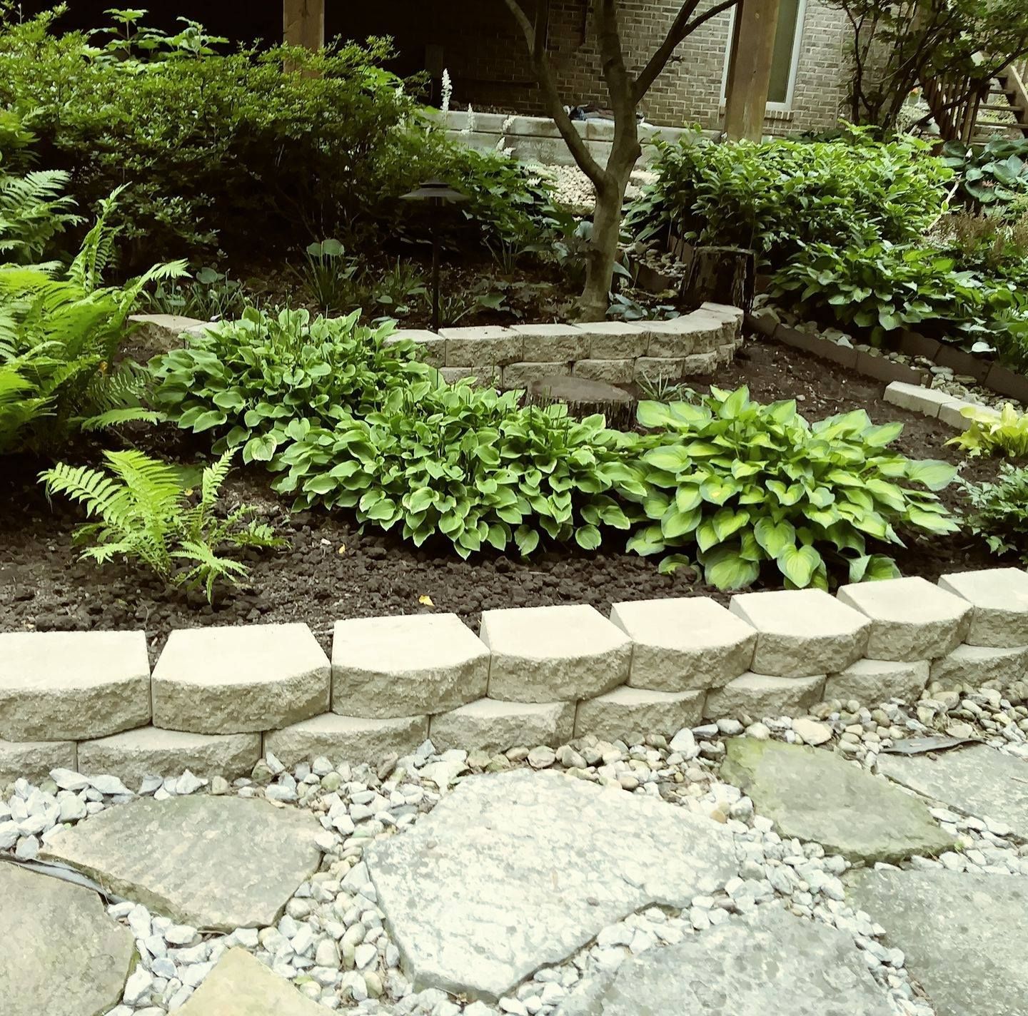 A garden with a stone patio in the foreground, raised garden beds with hostas and ferns, and a stone retaining wall.