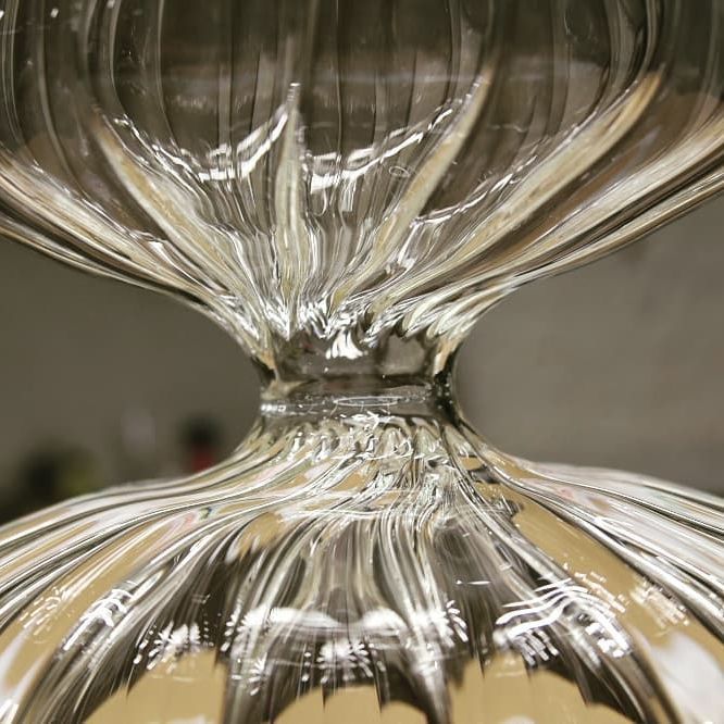 Primo piano di un oggetto di vetro trasparente e tondeggiante, con una vita schiacciata, che mostra creste verticali e riflessi.