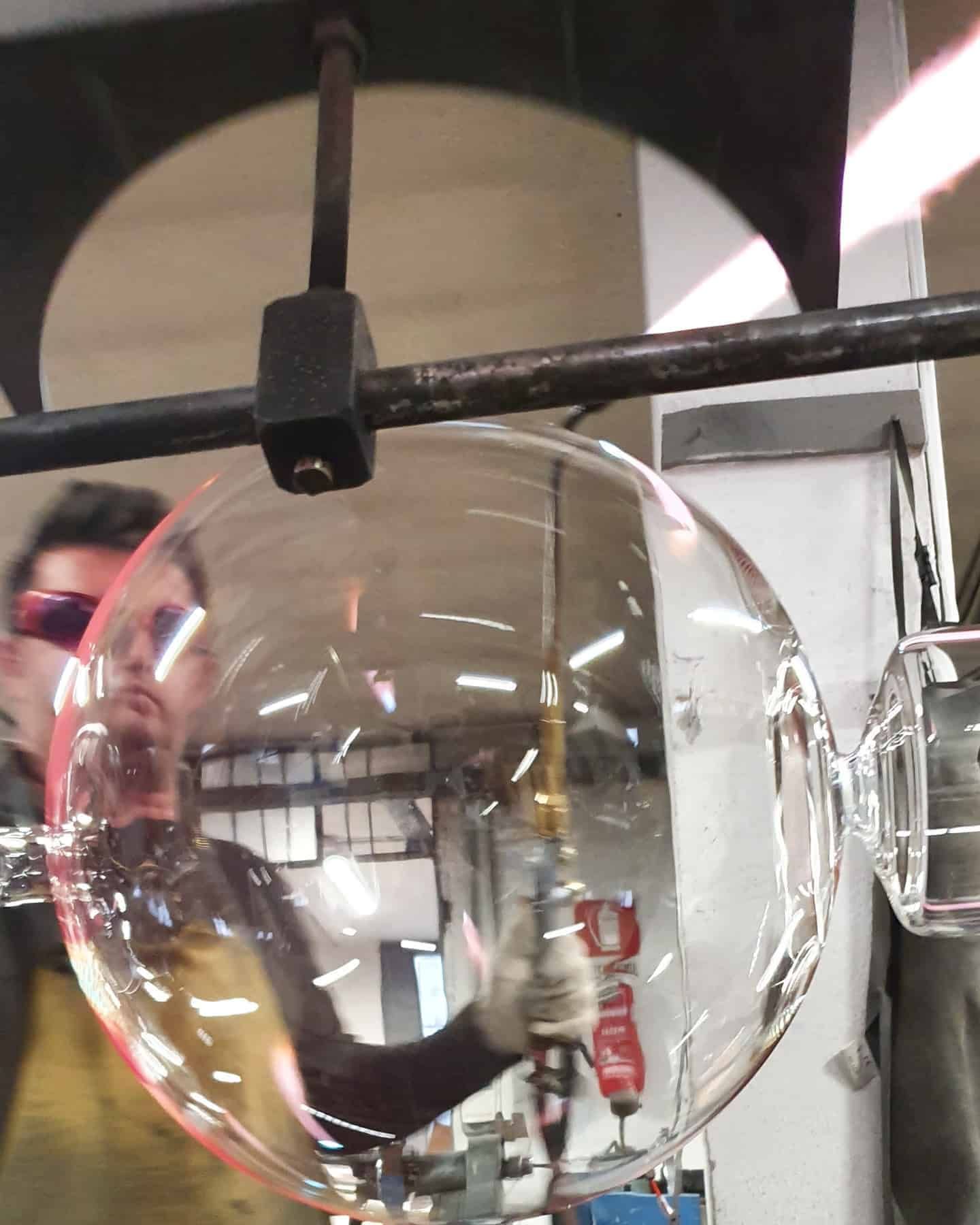 Soffiatore di vetro che modella una grande sfera di vetro trasparente con gli strumenti. Ambiente di laboratorio, persona che indossa occhiali protettivi.