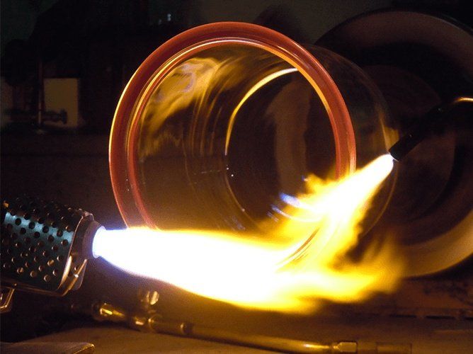 La fiamma di una torcia riscalda un anello di metallo bordato di rosso; l'ambientazione è scura, industriale.