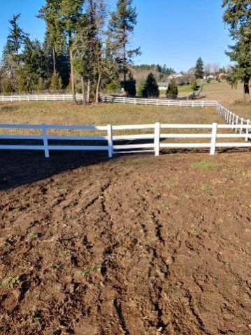 White fence around a brown dirt field with trees and a blue sky in the background