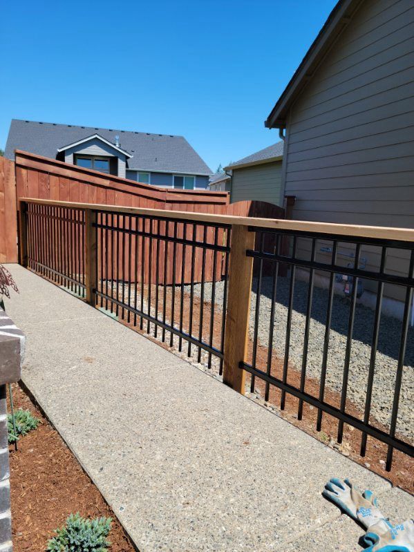Narrow side yard with gravel path, wooden fence, black metal railing, and house walls under a blue sky