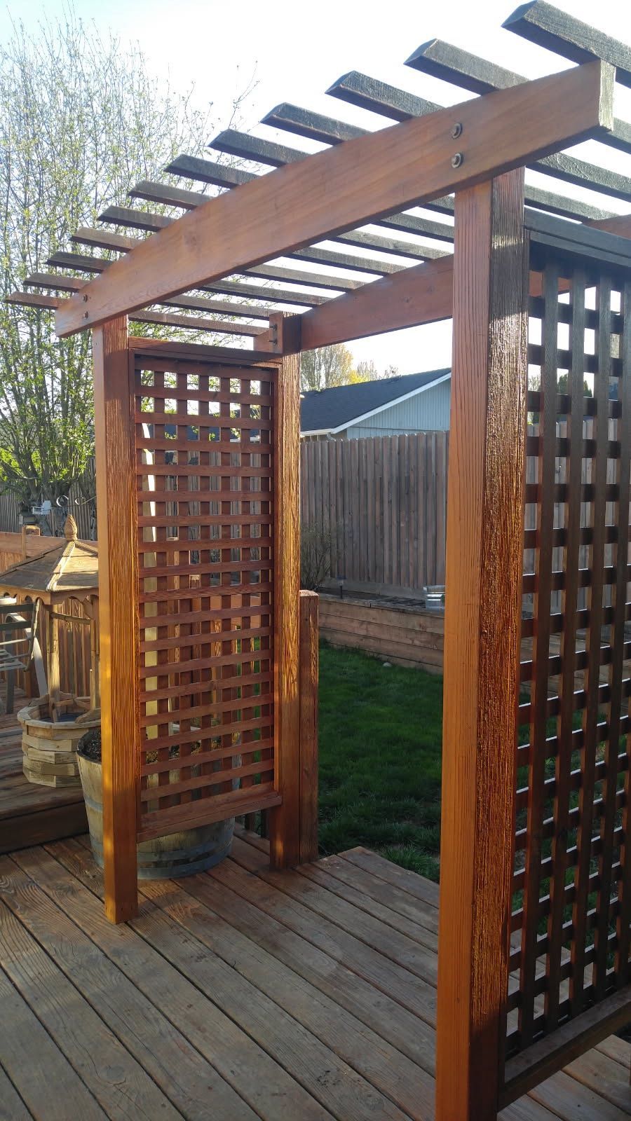 Wooden garden arbor with lattice sides on a deck, framed by a fence and greenery.