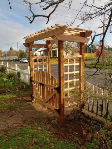 Wooden garden arbor gate beside a white picket fence and roadside, with bare trees in the background