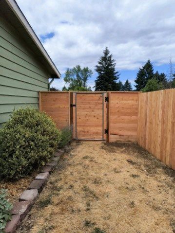 Narrow backyard with wooden gate, side fence, and green house wall under cloudy sky