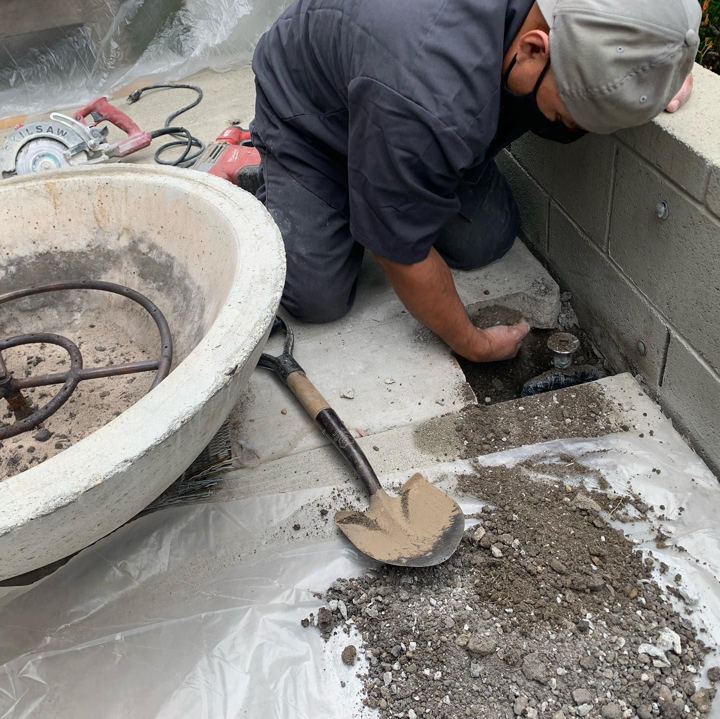 A person kneels on a patio, working on a drain, shovel nearby. A concrete fire pit sits to the left.