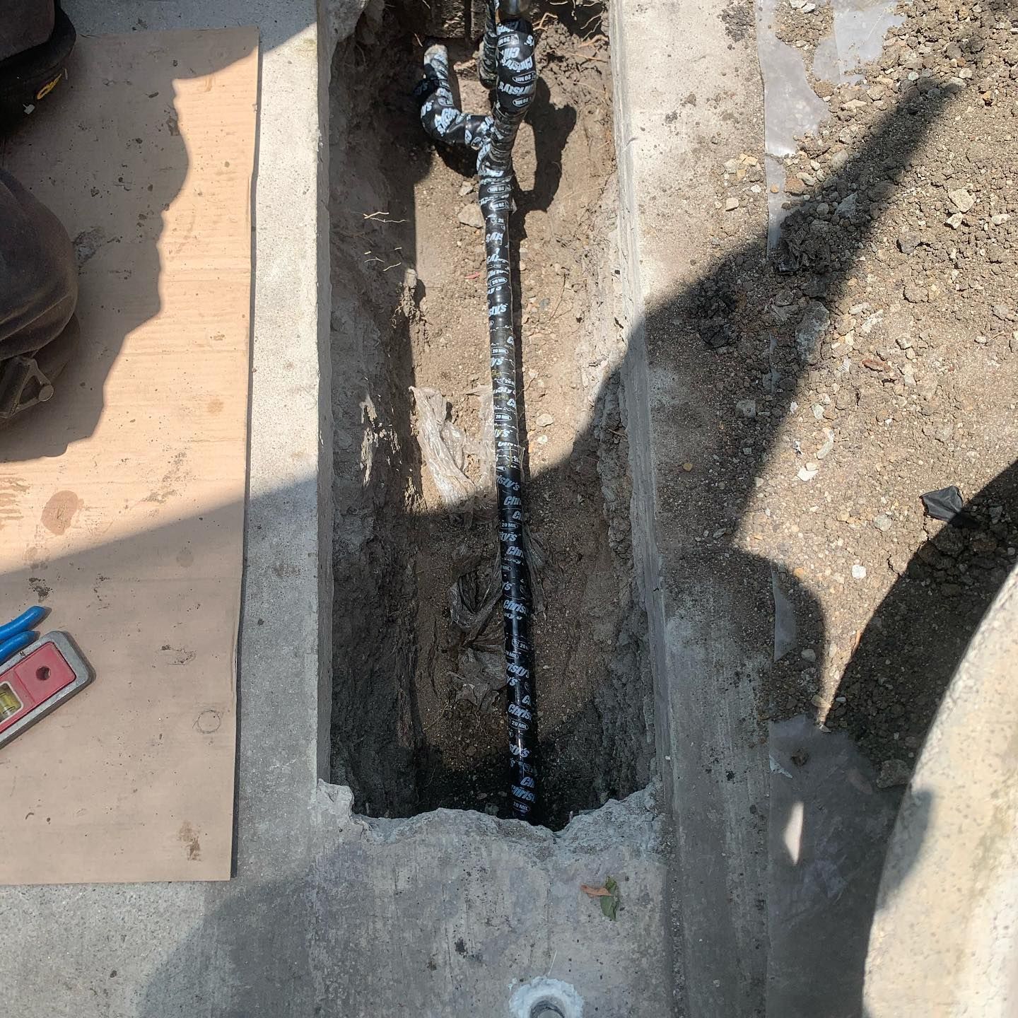 A narrow trench in concrete revealing a section of black pipe with silver fittings.