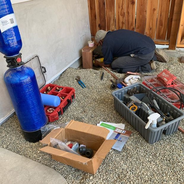 A person repairs plumbing outdoors next to a water filtration system. Tools and parts are scattered on a gravel surface.