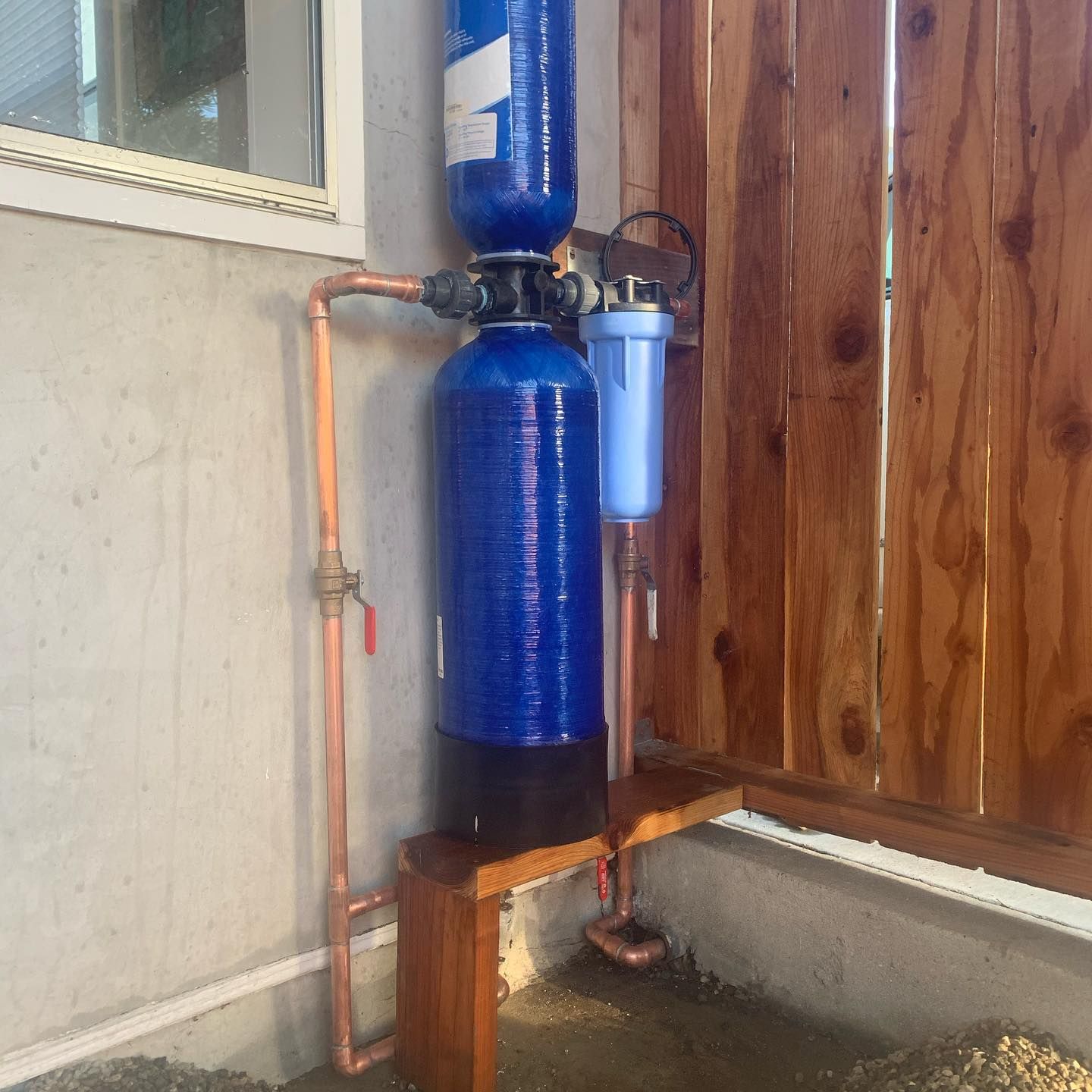 Water filtration system, comprised of blue tanks and filters, installed outdoors next to a wooden fence and concrete wall.
