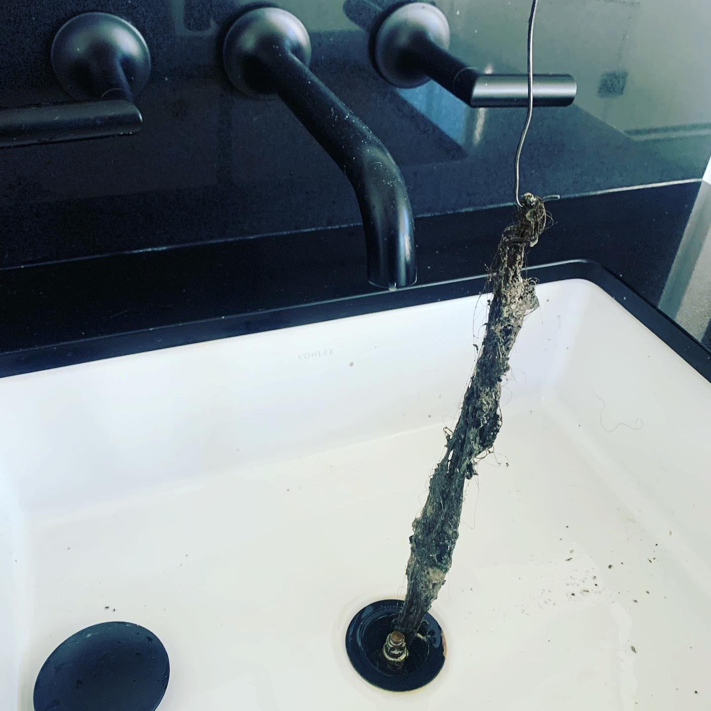 A clogged sink shows a tangled mass of hair and debris pulled out of the drain by a wire tool. Black faucet and white sink.
