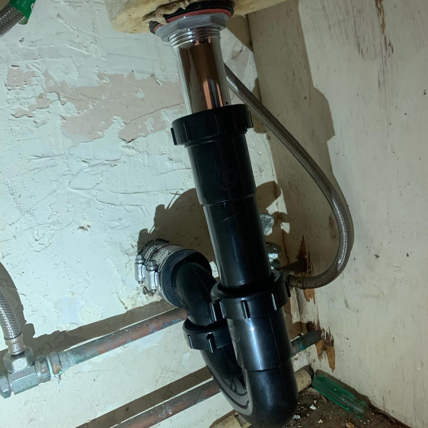 Under-sink plumbing with black pipe, chrome drain, and a flexible water supply line. The pipes are against a beige wall.