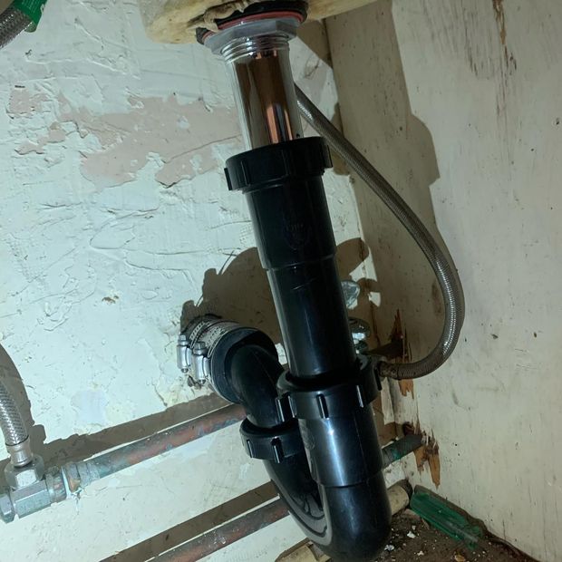 Under-sink plumbing with black pipe, chrome drain, and a flexible water supply line. The pipes are against a beige wall.