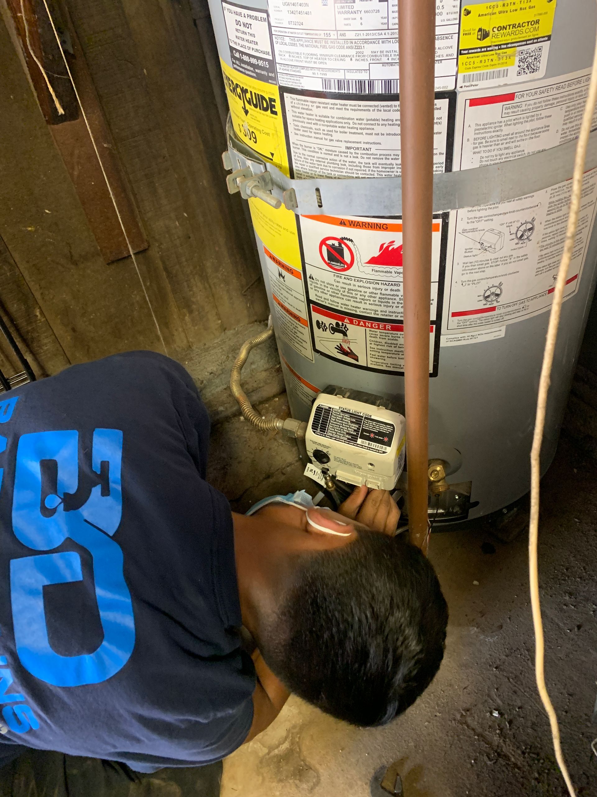 A person in a blue shirt working on a water heater in a basement. The person is lying on the floor.