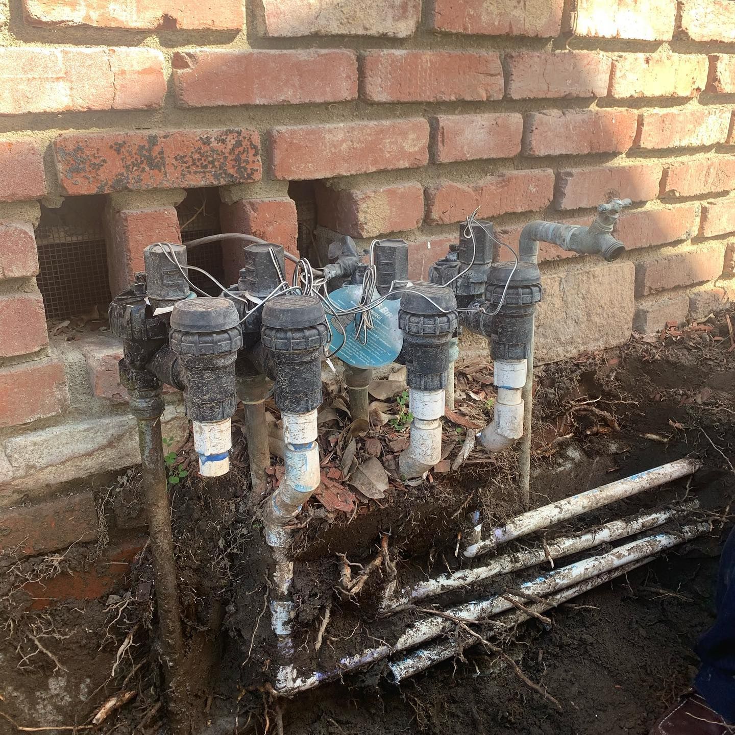 Sprinkler system valves and pipes installed against a brick wall in an outdoor setting, partially buried in soil.