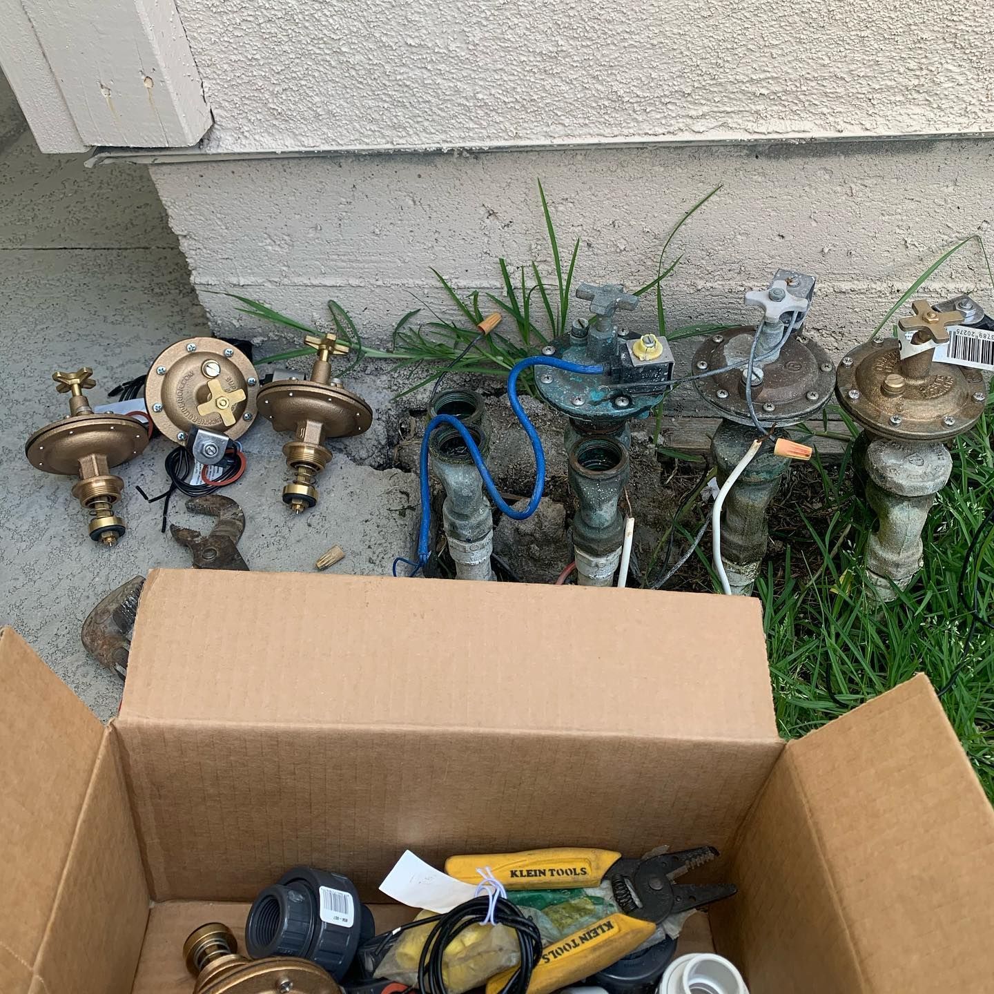 Cardboard box with sprinkler heads and parts, next to a concrete foundation. Some sprinkler heads are set in the ground.