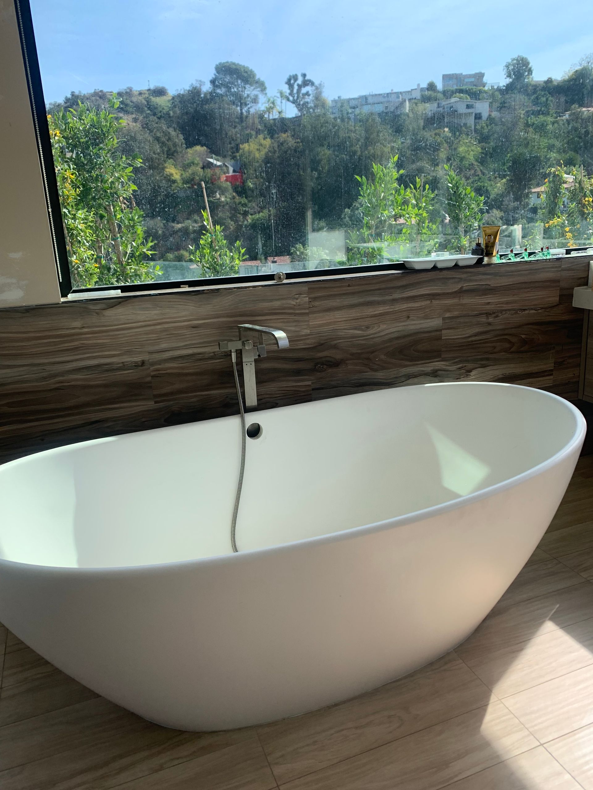 An oval white bathtub sits in front of a large window overlooking a green hillside. Sunlight streams in.