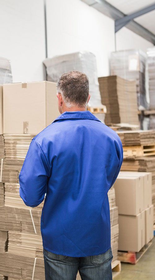 A man in a blue jacket is standing in a warehouse looking at boxes.